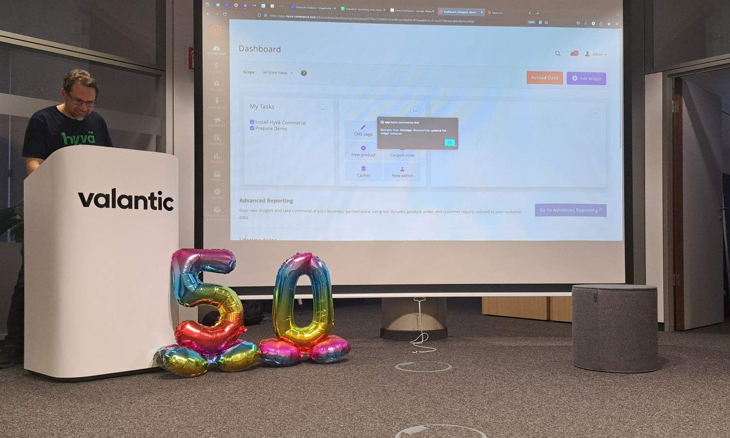 Die Präsentation wird per Beamer an die Leinwand geworfen, in der unteren rechten Ecke sind zwei regenborgenfarbene Ballons platziert, die zusammen die Zahl 50 zeigen.