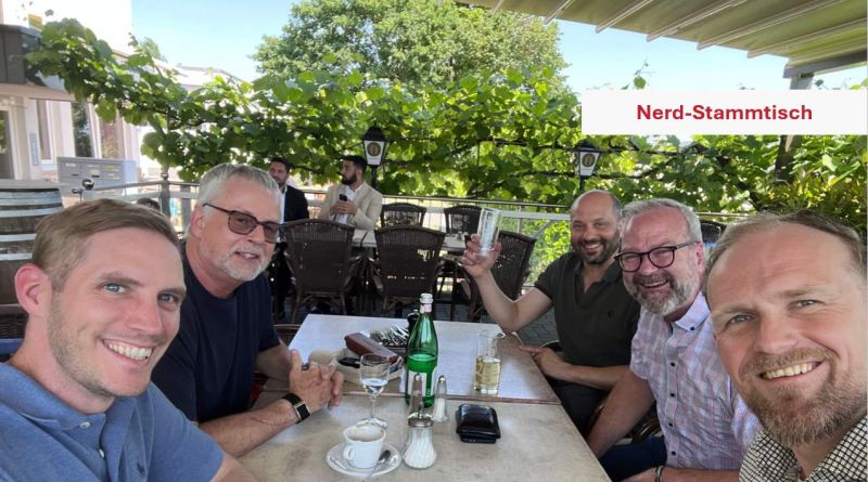 Fünf Männer sitzen an einem Tisch auf einer begrünten Terrasse; oben rechts steht „Nerd-Stammtisch“.